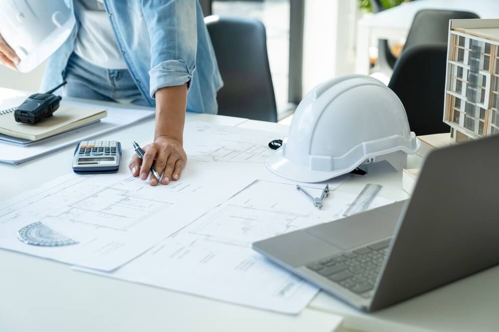 construction companies_Engineer or architect working at desk with building model and equipments in office workplace using laptop, thinking, planning and discussing about blueprints of building construction project.