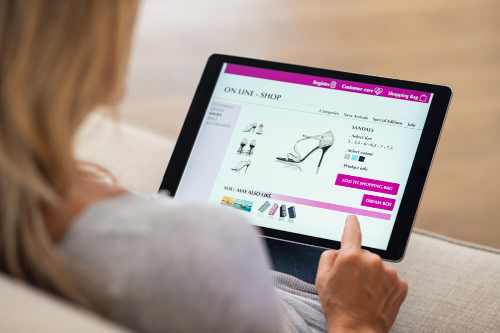 eCommerce_Closeup of woman doing online shopping on digital tablet at home. Rear view of woman hand touching screen while selecting shoes on ecommerce portal. Lady use e-commerce webshop to buy shoes.