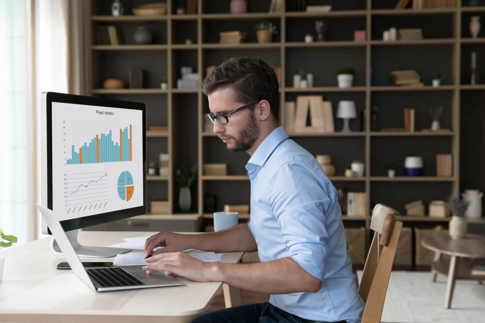 analytics_Focused young freelance business man analyzing financial data on desktop large display, working on marketing chart, sales report, using modern Internet technology for job, typing on laptop