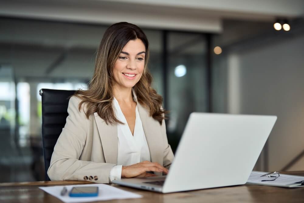 business owners_Smiling busy mature middle aged professional business woman manager executive wearing suit looking at laptop computer technology in office working on digital project sitting at desk.