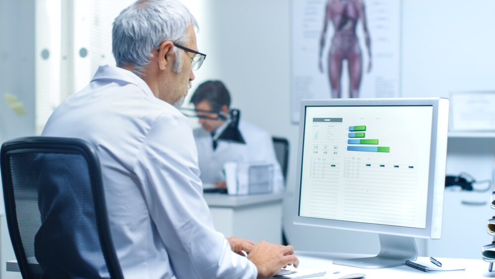 healthcare analytics_Senior Male Laboratory Researcher Working with Graphs on His Personal Computer. His Assistant Works at His Desk in the Background.