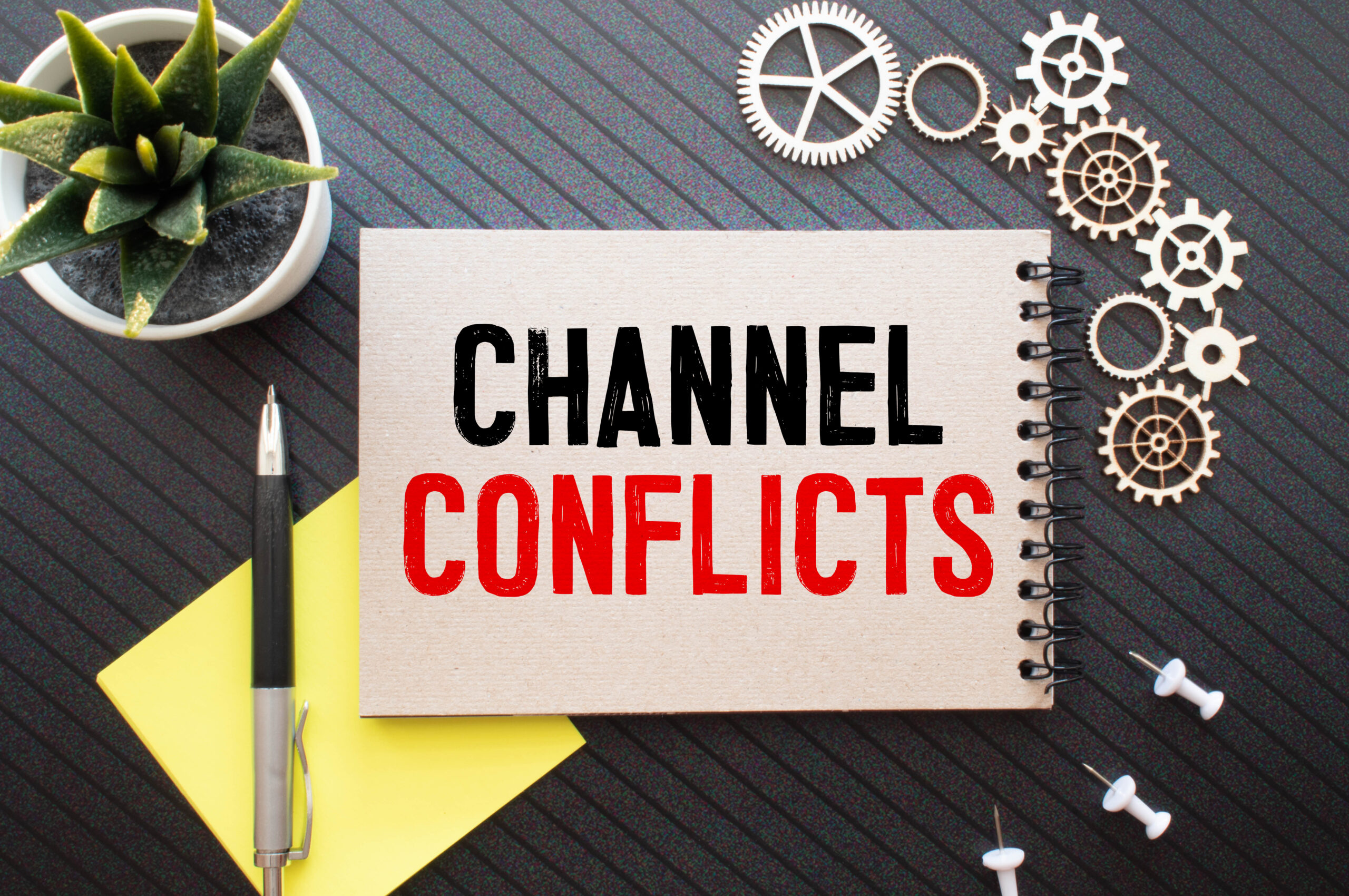 Notepad with the words "channel conflicts" in red and black is next to wooden gears, a pen, sticky notes, a succulent, thumbtacks to represent a fractional CMO consultant for channel conflict resolution in Chicago