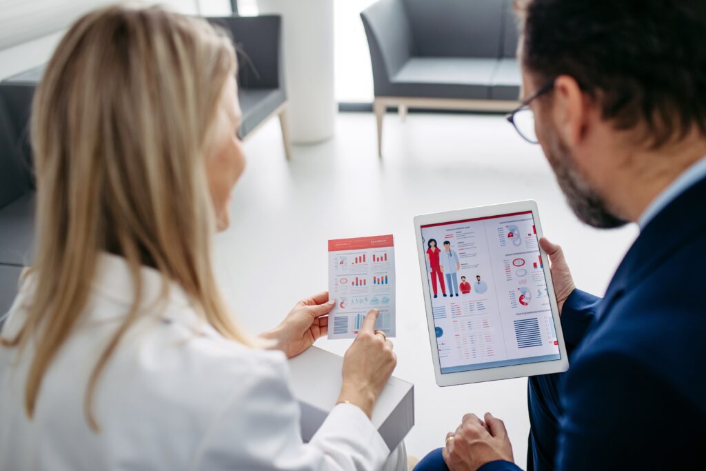 A doctor in a lab coat with long blond hair and a businessman in glasses review a pamphlet and tablet covering marketing challenges and tips for doctors.