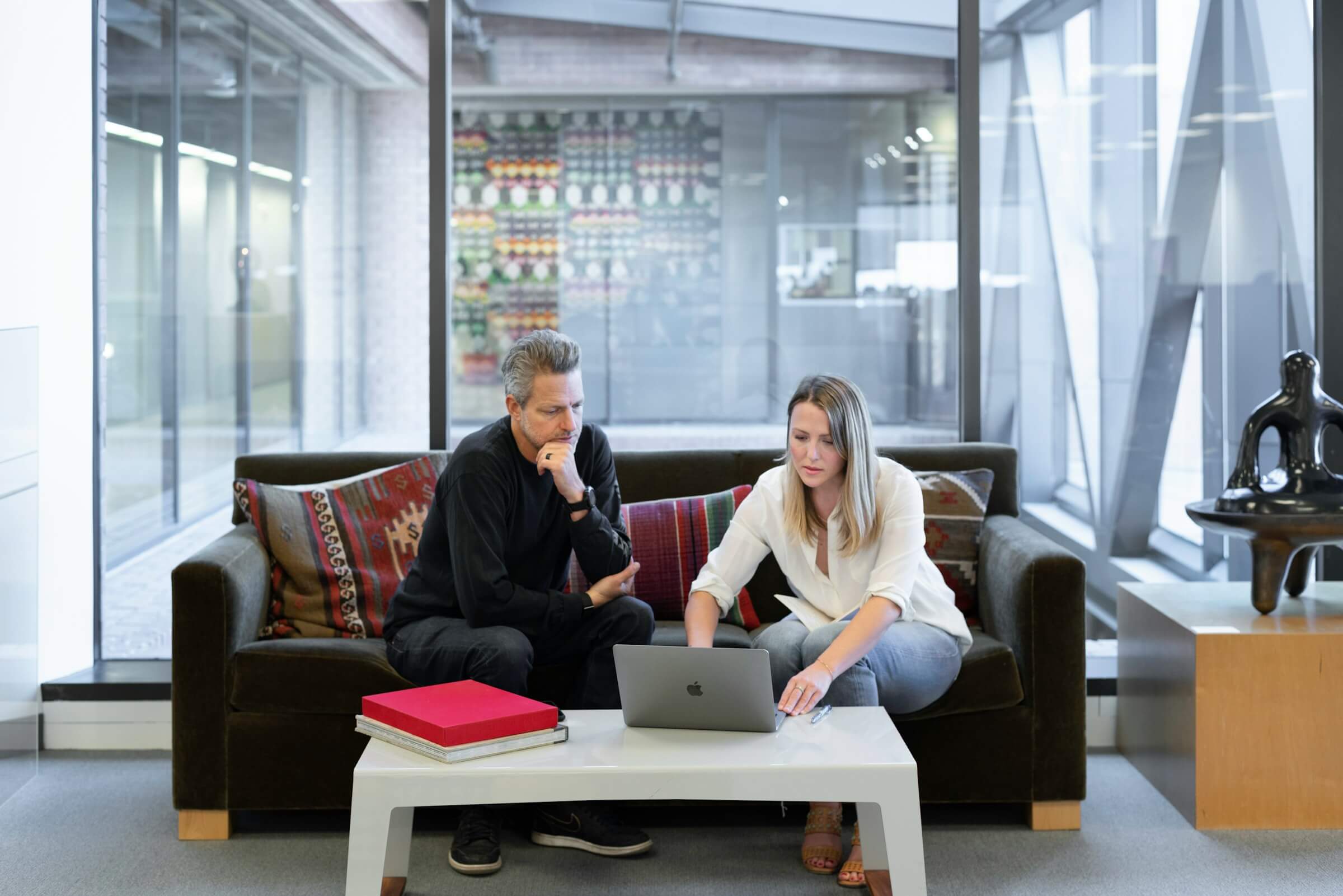 Businessman and fractional marketing director sit on a gray office couch and review winning strategy on a laptop