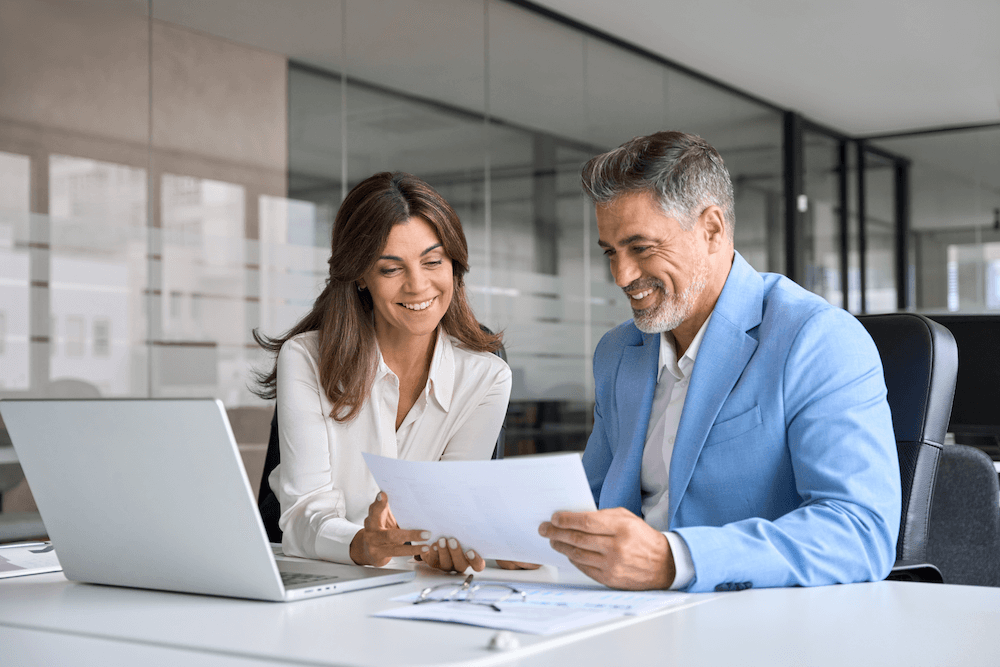 A fractional marketing director shows a client a new strategy at a desk on a laptop and paper sheets