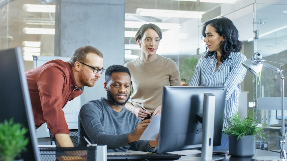 Group of four B2B Marketing Consultants gather in front of a desktop computer in an office to review a campaign