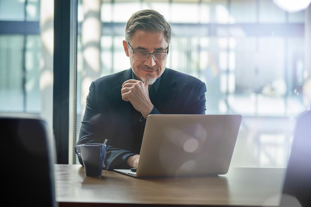 a B2B marketing consultant with gray hair and glasses types at his computer next to a black coffee mug