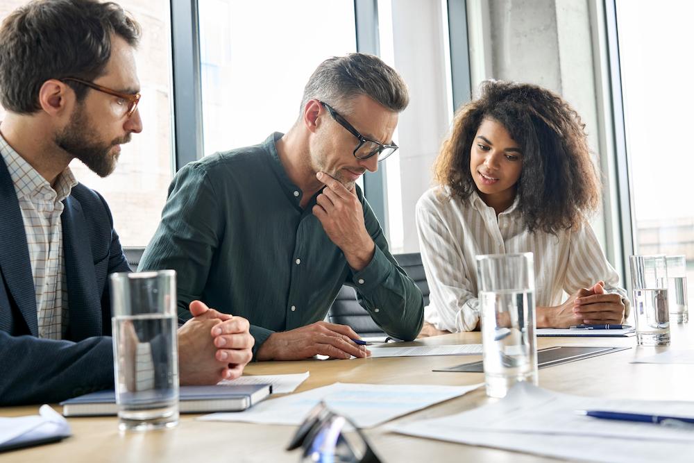 A marketing strategy consultant sits between two team members as they review their printed strategies. On the desk are glasses of water and loose papers.