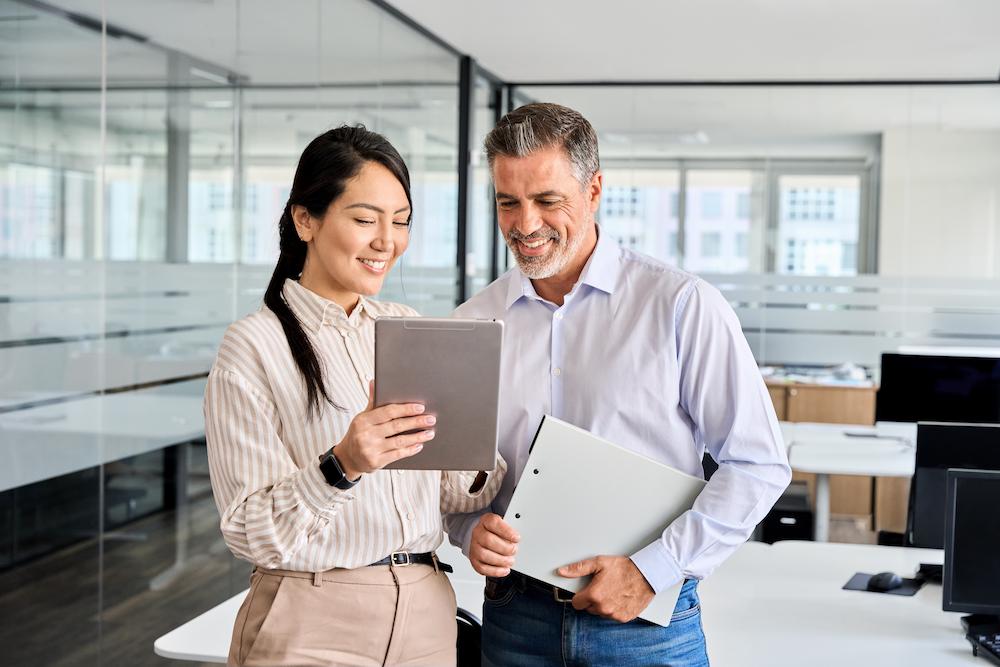 In an office, one marketing strategy consultant holds up a tablet. The other strategist looks on and holds a notebook.